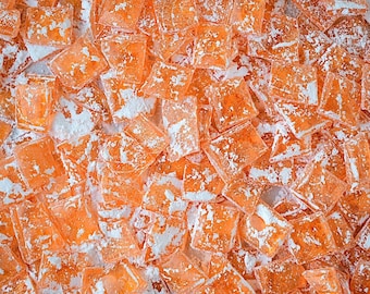 May include: Overhead shot of a pile of orange, square-shaped candies dusted with white powder. The candies are translucent, and the white powder creates a textured appearance. The candies are scattered across the frame.