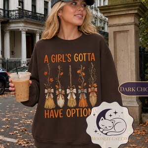 May include: A dark chocolate brown crewneck sweatshirt featuring the orange text A GIRL'S GOTTA HAVE OPTIONS above a graphic of five distinct witch brooms decorated with bows and autumn leaves, paired with a dark grey baseball cap.