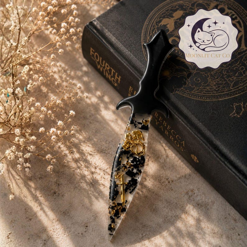 May include: A decorative dagger with a black handle and a clear blade embedded with gold accents and black flecks. The dagger is resting on a textured surface next to a book. The dagger has a unique design.