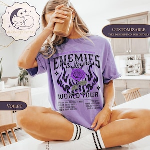 May include: A lilac t-shirt with the text "Enemies to Lovers World Tour" and a purple rose graphic. The shirt also features a list of dates and names. The shirt is part of the "Moonlit Cat Co" collection.