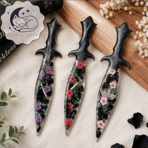 May include: Three decorative daggers with black handles and clear blades filled with flowers. The flowers are purple, red, and pink. Each dagger has a different silver emblem: a sword, a crown, and a cross.