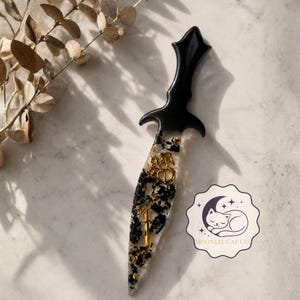 May include: A decorative dagger with a black handle and a clear blade filled with black and gold accents. The handle has a gothic design. The blade features gold embellishments, including a cross and a decorative emblem. The dagger is a unique art piece.