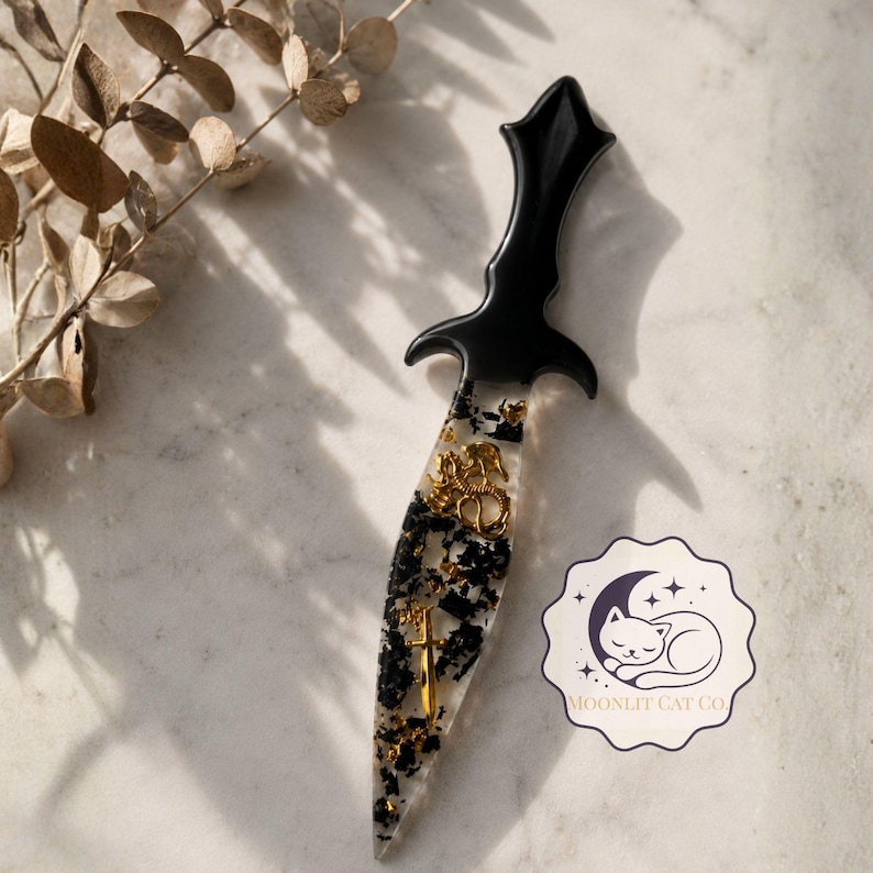 May include: A decorative dagger with a black handle and a clear blade filled with black and gold accents. The handle has a gothic design. The blade features gold embellishments, including a cross and a decorative emblem. The dagger is a unique art piece.
