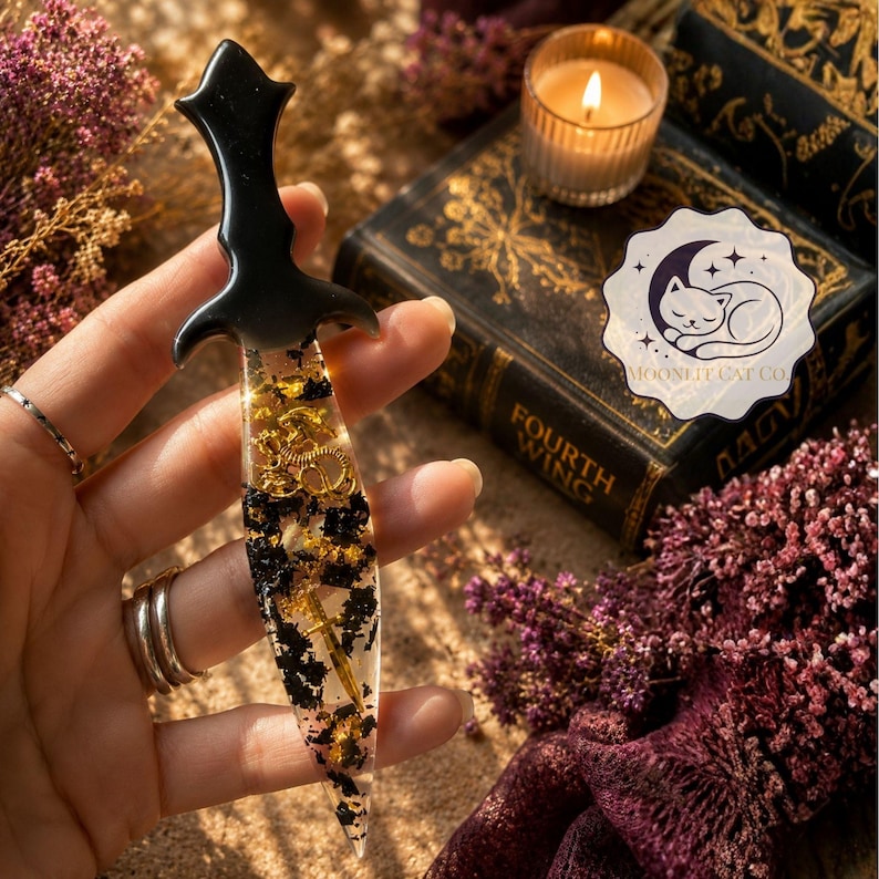 May include: A black resin dagger-shaped bookmark with gold and black accents. The bookmark features a decorative handle and is filled with gold-colored gears and black floral elements. The product is from Moonlit Cat Co.