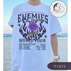 May include: White t-shirt with a graphic design. The design includes the text "Enemies to Lovers World Tour" and a purple rose. The shirt also lists tour dates and featured artists. A Moonlit Cat Co. logo is visible.