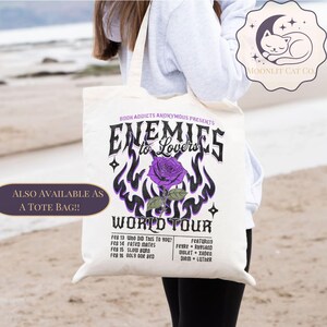 May include: A cream tote bag with the words "Enemies to Lovers World Tour" and a purple rose design. The bag also lists dates and names. The bag is being held by a person.