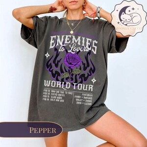 May include: A pepper-coloured graphic tee with the text "Enemies to Lovers World Tour", a purple rose, and flame design. The tee lists tour dates and featured artists. A Moonlit Cat Co. logo is visible.