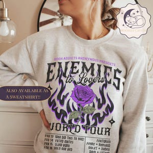 May include: Pale grey sweatshirt with a graphic design. The design features the text "Enemies to Lovers" and a purple rose. The design includes the words "World Tour" and a list of dates. The text "Also available as a sweatshirt!!" is also visible.
