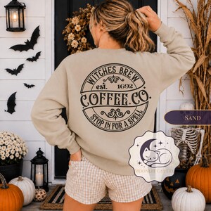 May include: A sand-colored long-sleeve sweatshirt featuring a black circular graphic on the back with the text Witches Brew est. 1692 Coffee Co. Stop In For A Spell. The design includes decorative flourishes and a small logo with a sleeping cat.