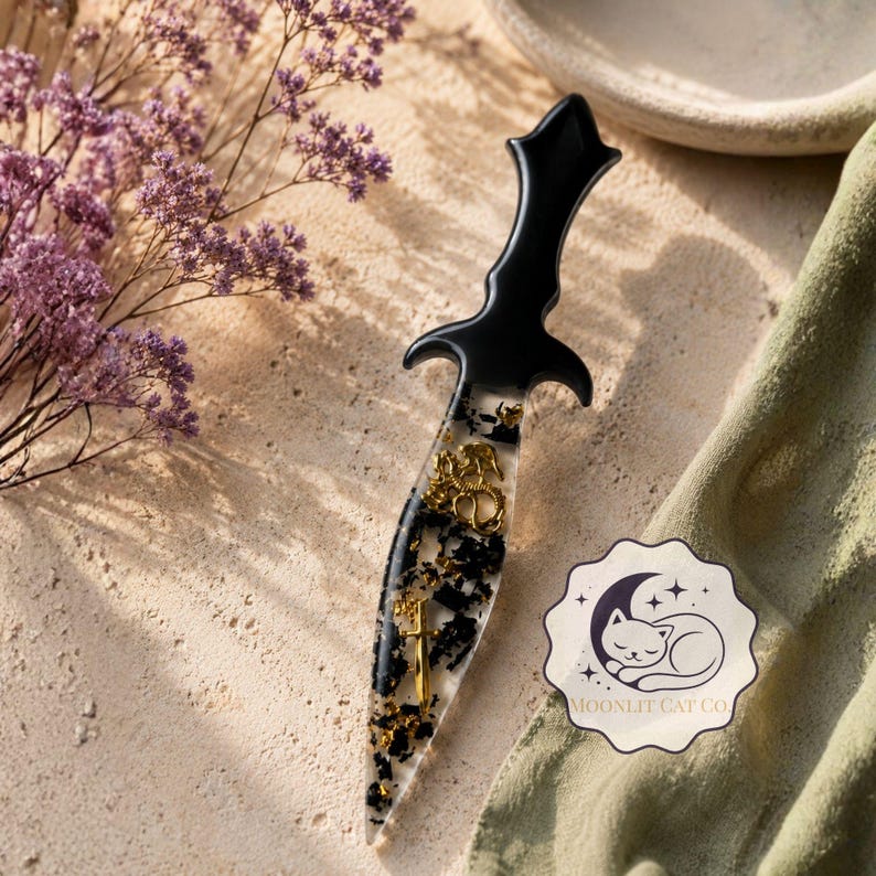May include: A decorative dagger with a black handle and a clear blade filled with black and gold accents. The dagger features gold embellishments and is set on a textured surface. A sticker with a sleeping cat is in the image.