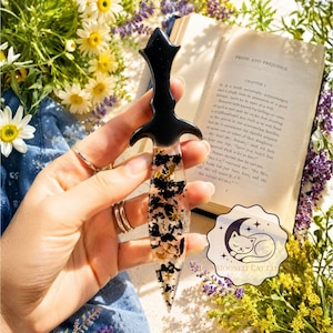 May include: A black and clear resin dagger-shaped bookmark with gold and black inclusions. The bookmark has a decorative handle and is held in front of an open book with the text "Pride and Prejudice".