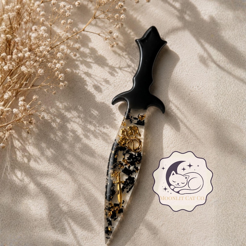 May include: A decorative dagger with a black handle and a clear blade filled with gold flakes and black accents. The blade features gold embellishments, including a cross and a crest. A logo for Moonlit Cat Co. is visible.