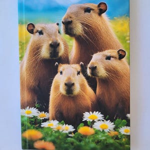 Könnte beinhalten: Ein Notizbuch mit einem lebendigen Bild einer Capybara-Familie in einem Gänseblümchenfeld. Das Cover zeigt vier braune Capybaras mit weichem Fell, vor dem Hintergrund von grünem Gras, gelben Löwenzahnpflanzen und blauem Himmel.