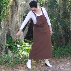 May include: A woman wearing a white long-sleeved shirt and a brown overall dress with straps. She is standing in a forest with moss hanging from the trees. She is wearing white boots.
