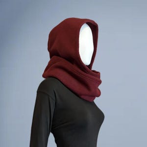 May include: A mannequin wearing a dark red hooded scarf draped around the neck and head, paired with a long-sleeved black shirt and black leggings. The ensemble features a solid color palette with a smooth texture on the shirt and leggings.