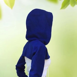 May include: A blue cropped hoodie with long sleeves and a hood. The hoodie is worn over a white top with a drawstring. The person is standing in a grassy field with a blurred background of green leaves.