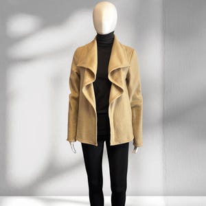 May include: A tan open-front jacket with a wide, draped collar and long sleeves, worn over a black mock-neck top and black leggings on a mannequin. The jacket features a soft, textured finish and a structured yet flowing silhouette.
