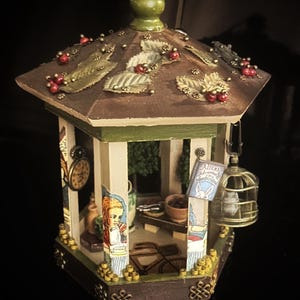 Könnte beinhalten: Ein Miniatur-Pavillon aus Holz mit einem grünen Dach, das mit Blättern und roten Beeren verziert ist. Der Pavillon hat ein weißes Interieur mit einem kleinen Tisch, einem Buch mit dem Titel "Alice's Adventures in Wonderland" und einem Vogelkäfig, der von der Decke hängt.