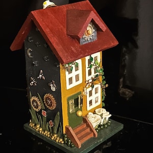 Puede incluir: Una casa de madera en miniatura con un techo rojo, paredes amarillas y molduras verdes. La casa tiene un pequeño porche con escalones y una puerta. La casa está decorada con flores, hojas y otros detalles.