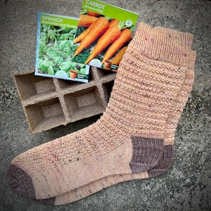Könnte beinhalten: Ein Paar gestrickte Socken in Beige und Braun mit einem gesprenkelten Muster. Die Socken liegen auf einer braunen Pappe mit Saatgutpaketen für Spinat und Karotten.
