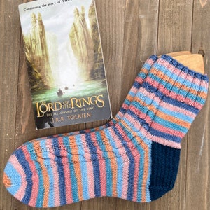 Puede incluir: Un par de calcetines de punto a rayas con un patrón de trenza. Los calcetines son en tonos de azul, rosa, naranja y turquesa. Los calcetines están sobre una superficie de madera junto a una copia de "El Señor de los Anillos: La Comunidad del Anillo" de J.R.R. Tolkien.