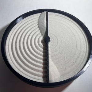 May include: A black circular sand timer with a divider in the middle. The sand is white and creates a pattern of concentric circles on one side of the divider.
