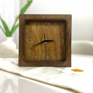 Op de afbeelding: Een vierkante houten klok met een donkerbruine afwerking en zichtbare houtnerf. De wijzerplaat is voorzien van uitgesneden cijfers op de 12, 3, 6 en 9 uur posities, met eenvoudige streepjes voor de overige uren en drie dunne zwarte wijzers.