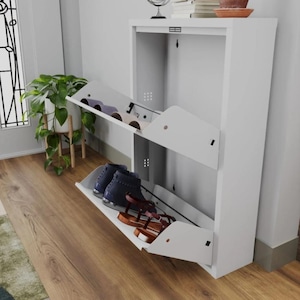 Op de afbeelding: Een witte schoenenkast met twee neerklapbare compartimenten, elk met meerdere paar schoenen. De kast staat tegen een muur, met een plant en decoratieve items erbovenop. De schoenen omvatten sandalen en laarzen.
