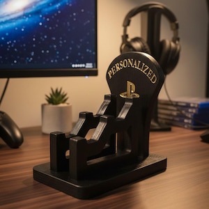 以下が含まれることがあります： 「PERSONALIZED」の金色の文字が入った黒い木製のゲームコントローラー用スタンド。スタンドには金のPlayStationロゴがあり、コントローラーを保持するように設計されています。ヘッドフォンのペアが背景のスタンドに置かれています。