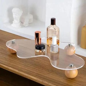 Puede incluir: Una bandeja de acrílico transparente con un diseño ondulado, sostenida por cuatro pequeñas esferas de madera. La bandeja exhibe productos de belleza, incluyendo un frasco de perfume, lápiz labial y esmalte de uñas, sobre una superficie de madera. El fondo muestra una estantería blanca con una estatua.