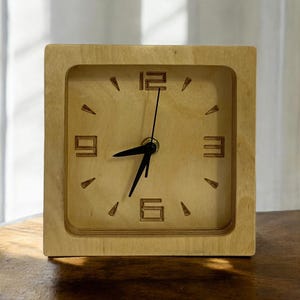 Op de afbeelding: Vierkante houten klok met een lichte houten afwerking. De wijzerplaat heeft zwarte wijzers en cijfers, met driehoekige markeringen. De cijfers zijn in een eenvoudig, modern lettertype. De klok staat op een houten oppervlak.