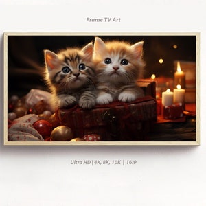 Puede incluir: Dos gatitos adorables, uno marrón atigrado y otro blanco con marcas naranjas, están sentados en una caja roja con una tapa de madera. Los gatitos miran a la cámara con expresiones curiosas. La caja está rodeada de decoraciones navideñas, incluyendo adornos rojos y dorados, y velas.