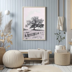 May include: A black and white photograph of a lone tree in a field with a wooden fence. The photo is framed in a light wood frame and hanging on a light blue wall.