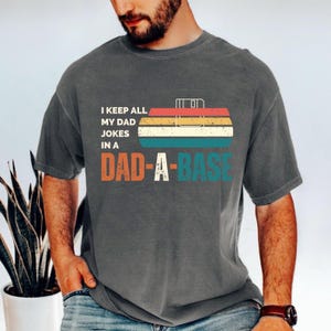 May include: Dark gray t-shirt with the text "I KEEP ALL MY DAD JOKES IN A DAD-A-BASE" in orange, teal, and white. The design includes a retro-style toolbox graphic with red, yellow, and teal stripes.