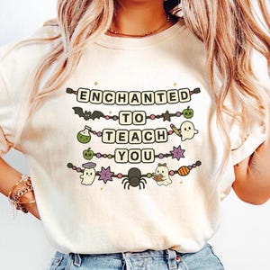 May include: Cream-colored t-shirt with the words "ENCHANTED TO TEACH YOU" in black block letters. The design includes Halloween-themed illustrations like a bat, potion bottle, ghost, and spider. The shirt is a casual, festive garment.