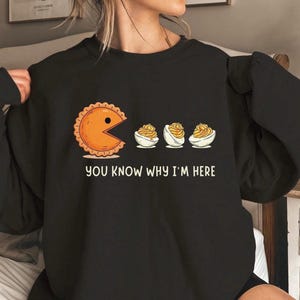 May include: Black sweatshirt with a graphic featuring a pie-shaped character eating deviled eggs, inspired by the Pac-Man game. The text "YOU KNOW WHY I'M HERE" is printed below the graphic. The design is in shades of orange, white, and yellow.
