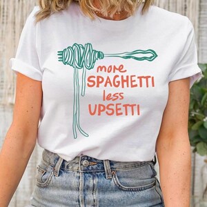 May include: White t-shirt with a green illustration of spaghetti on a fork and the text "more spaghetti less upsetti" in red and black.