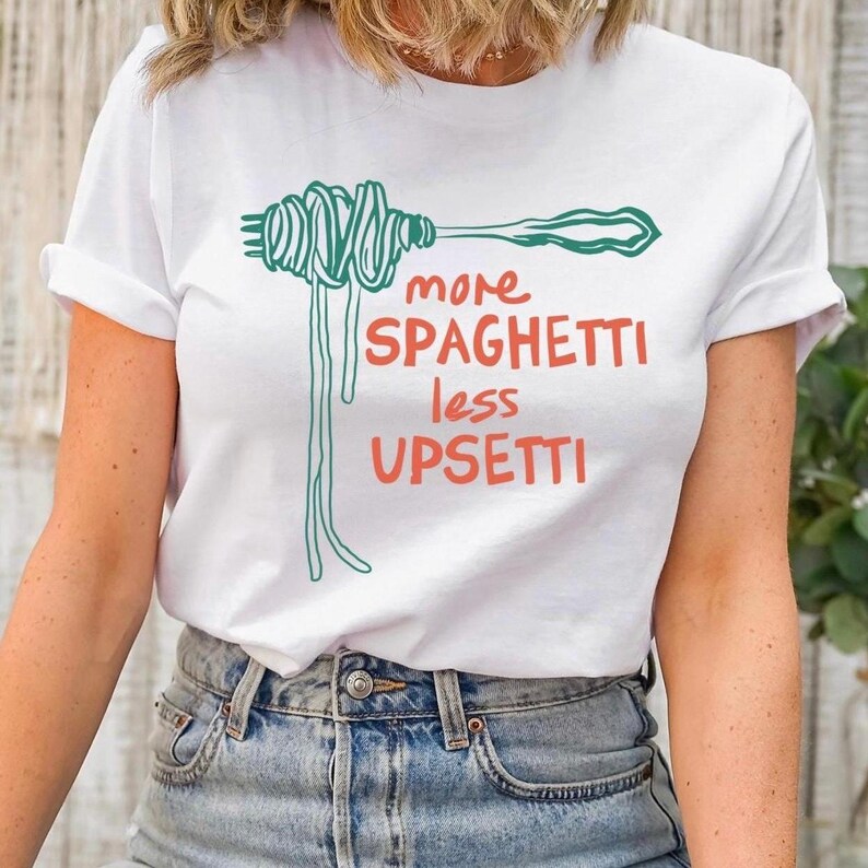 May include: White t-shirt with a green illustration of spaghetti on a fork and the text "more spaghetti less upsetti" in red and black.