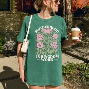 May include: A green t-shirt with the words "MOTHERHOOD IS KINGDOM WORK" in white and a floral design in pink and green. The shirt also includes the Bible verse Proverbs 31:10.
