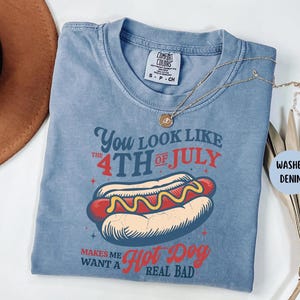 K&ouml;nnte beinhalten: Blaues Comfort Colors T-Shirt mit dem Text "You look like the 4th of July makes me want a hot dog real bad." Die Grafik zeigt einen Hotdog mit Senf. Eine goldene Halskette liegt &uuml;ber dem Shirt.