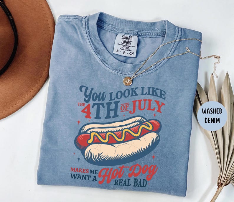 K&ouml;nnte beinhalten: Blaues Comfort Colors T-Shirt mit dem Text "You look like the 4th of July makes me want a hot dog real bad." Die Grafik zeigt einen Hotdog mit Senf. Eine goldene Halskette liegt &uuml;ber dem Shirt.