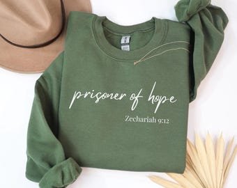 May include: An olive green sweatshirt with the words "prisoner of hope" in a cursive font, with "Zechariah 9:12" below. A gold necklace and a tan hat are visible in the background. The sweatshirt is made of a soft material.