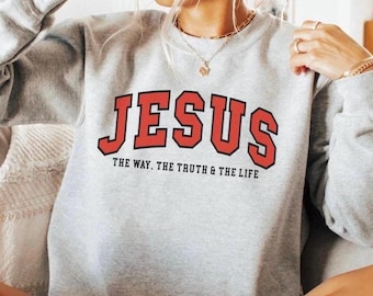 May include: A light gray sweatshirt with the word "JESUS" in large red block letters outlined in black. Below the word "JESUS" is the text "THE WAY. THE TRUTH & THE LIFE" in a smaller font. The sweatshirt is a crew neck style.