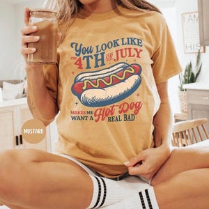 K&ouml;nnte beinhalten: Senffarbenes T-Shirt mit einem Hotdog-Motiv und dem Text "You look like the 4th of July makes me want a hot dog real bad." Das T-Shirt hat einen l&auml;ssigen Stil.
