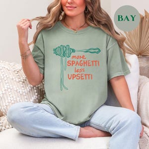 May include: A woman wearing a sage green t-shirt with the text "more spaghetti less upsetti" and a graphic of spaghetti noodles. She is sitting on a white couch with a white throw pillow.