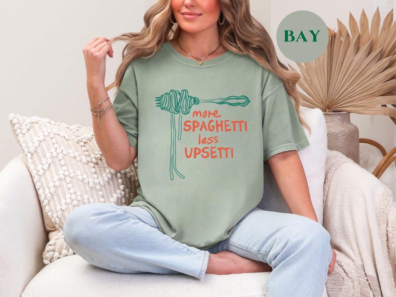 May include: A woman wearing a sage green t-shirt with the text "more spaghetti less upsetti" and a graphic of spaghetti noodles. She is sitting on a white couch with a white throw pillow.