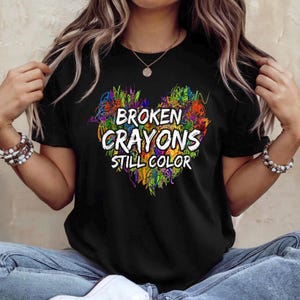May include: Black t-shirt with the text "BROKEN CRAYONS STILL COLOR" in white, surrounded by a colorful heart-shaped design. The design features a mix of vibrant colors, including green, orange, blue, and purple.