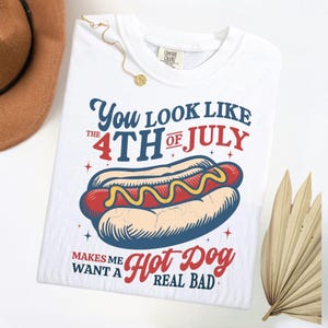 K&ouml;nnte beinhalten: Wei&szlig;es T-Shirt mit einem Hotdog-Motiv und dem Text "You look like the 4th of July makes me want a hot dog real bad." Der Hotdog ist rot mit gelbem Senf.