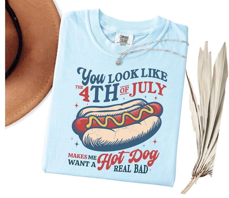 K&ouml;nnte beinhalten: Hellblaues T-Shirt mit einer Abbildung eines Hotdogs und dem Text "You look like the 4th of July makes me want a hot dog real bad." Eine silberne Halskette mit einem kleinen Anh&auml;nger liegt auf dem Shirt.