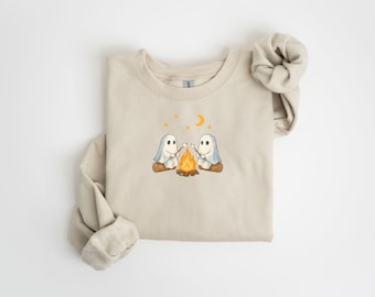 May include: Beige sweatshirt featuring an embroidered design of two cartoon ghosts roasting marshmallows over a campfire. The design includes a crescent moon and stars. The sleeves are rolled up. A cozy, casual top for Halloween or fall.